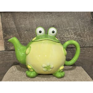 May include: A whimsical ceramic teapot shaped like a frog. The teapot is primarily light green and yellow, with large, cartoonish eyes on the lid. The handle and spout are curved, and the frog's feet are visible at the base.