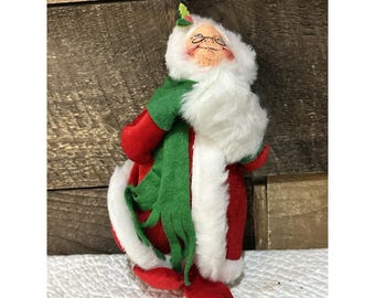 Puede incluir: Una figura de Papá Noel con abrigo rojo con ribete de piel sintética blanca, una bufanda de fieltro verde y una cara pintada con gafas. La figura tiene una barba de piel sintética blanca y un pequeño adorno de hoja de acebo verde.