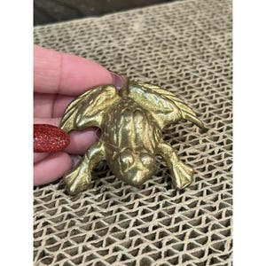 May include: A brass frog figurine with outstretched wings and detailed features. The frog is in a crouching position, with its legs spread and eyes forward. The figurine is a golden color.