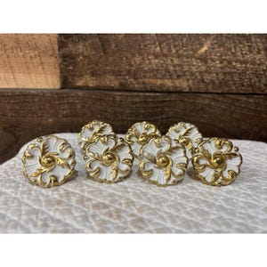 May include: A set of seven circular buttons featuring a white base with raised gold-colored floral and leaf patterns. Each button has a central gold-colored dot and a scalloped edge, showcasing a detailed metallic finish against the white enamel.
