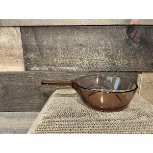 Puede incluir: Una taza medidora de vidrio transparente de color ámbar con un asa. La taza es redonda con un pico vertedor y está sobre una superficie texturizada. El fondo es una pared de madera.