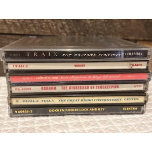以下が含まれることがあります： CDケースが6つ積み重なった写真。Train、Winger、Collective Soul、Bonham、Tesla、DokkenなどのアーティストのCDが入っています。ケースは透明なプラスチック製で、アルバムタイトルとアーティスト名が記載されています。