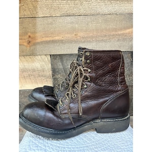 Puede incluir: Un par de botas de trabajo de cuero marrón oscuro con cierre de cordones con ojales y ganchos metálicos, punta redondeada, suela gruesa de goma negra y un panel decorativo de flecos de cuero sobre la base de los cordones.
