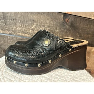 May include: Black leather clogs with a decorative design and a wooden platform heel. The clogs feature a scalloped edge, metal studs, and a decorative metal button. The platform heel measures approximately 3 inches.