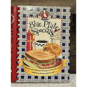 Puede incluir: Un libro de cocina titulado "Blue Plate Specials" con encuadernación en espiral roja. La portada presenta una ilustración de estilo restaurante de un sándwich, aros de cebolla, café y un batido. El texto dice: "Recetas que a tu familia y amigos más les encantarán, inspiradas en restaurantes de costa a costa."