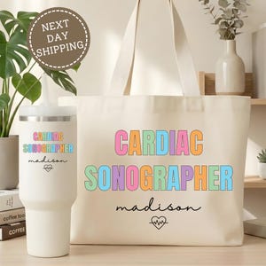 May include: A cream-colored tote bag and tumbler set. The tote bag and tumbler both feature the words "CARDIAC SONOGRAPHER" in colorful block letters, with the name "madison" and a heartbeat graphic below. The tumbler is approximately 10 inches tall.