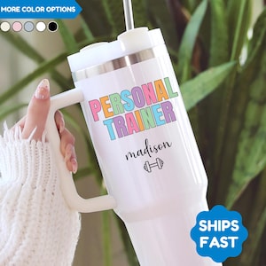 May include: White tumbler with a handle, featuring the text "PERSONAL TRAINER" in colorful letters. Below, the name "madison" is written in a script font, along with a dumbbell icon. The tumbler has a silver rim and a white lid.