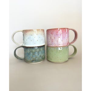 Peut inclure: Quatre tasses en céramique disposées en deux piles. La tasse en haut à gauche est bleu clair et crème, celle en bas à gauche est bleu foncé, celle en haut à droite est rose et celle en bas à droite est vert clair. Chaque tasse a des points en relief.