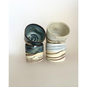 Peut inclure: Quatre tasses en céramique disposées en deux piles. La pile de gauche présente un intérieur et un bord émaillés bleu profond avec une base marbrée marron et crème. La pile de droite est crème avec des rayures horizontales marron et bleu clair.