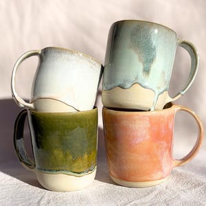 Peut inclure: Un ensemble de quatre tasses en céramique avec anses, empilées par deux. Chaque tasse présente une base beige non émaillée et une finition brillante en émail réactif dans des couleurs distinctes : blanc, vert sauge, vert olive et rose corail.