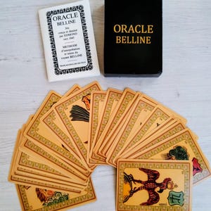 Peut inclure: Un jeu de cartes Oracle Belline, avec un étui noir et un livret blanc. Les cartes sont étalées, révélant des illustrations détaillées et des bordures décoratives. Le texte "ORACLE BELLINE" est présent sur l'étui et le livret. Les cartes sont beiges.