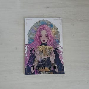 May include: A rectangular book titled Tarot Guide featuring an illustration of a person with long pink hair holding three gold tarot cards. The person wears a dark top with lace details, set against a circular stained glass window background.