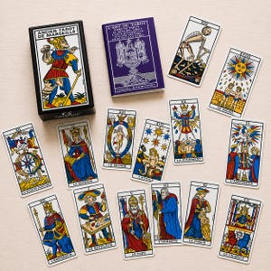 Puede incluir: Una baraja de cartas Ancien Tarot de Marseille con una caja negra, un libro de instrucciones morado titulado L'Art du Tarot de Alexandre Jodorowsky y catorce cartas de tarot individuales con ilustraciones coloridas y etiquetas de texto en francés.