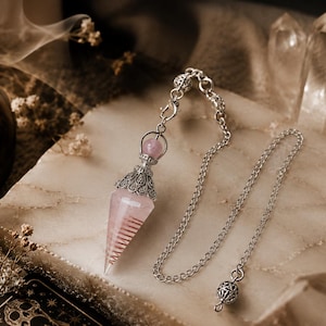 Könnte beinhalten: Ein spitz zulaufendes, blassrosa Kristallpendel mit einer silberfarbenen Metallkappe mit filigranen Details. Das Pendel ist an einer silberfarbenen Metallkette befestigt, die in Abständen kleine, dekorative kugelförmige Metallperlen aufweist.