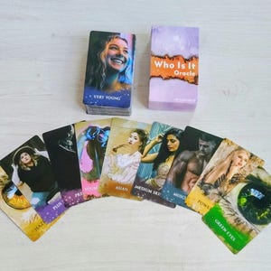 Puede incluir: Una baraja de cartas oráculo Who Is It con una caja morada y naranja. Las cartas presentan varios retratos y etiquetas de texto descriptivas que incluyen Very Young, Asian, Medium Skin, Muscular, Powerful y Green Eyes, dispuestas en abanico.