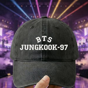 May include: A black baseball cap featuring the white text "BTS JUNGKOOK-97". The cap is held up in front of a concert stage with purple and blue lighting.