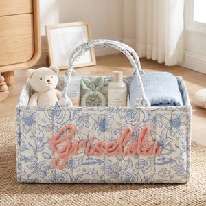 May include: A baby gift basket with a floral pattern in blue and white, containing a teddy bear, baby products, and a blanket. The name "Griselda" is spelled out in pink yarn on the front. The basket has a handle.