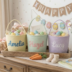 May include: Three pastel-colored Easter baskets with handles, each embroidered with a name: Amelia, Hazel, and Lucy. The baskets are filled with decorative eggs and sit on a wooden tray with carrot decorations and baby booties. A banner that says "Happy" hangs in the background.