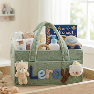 May include: A sage green baby gift basket filled with baby essentials. The basket includes a book, a crochet car, baby washcloths, a wooden teether, and a bottle of lotion. The name "Leroy" is spelled out on the front with fabric letters and a teddy bear.