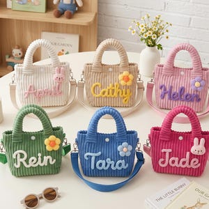 May include: Six corduroy tote bags in various colors: white, tan, pink, green, blue, and fuchsia. Each bag has a name embroidered on it, along with a decorative flower or bunny. The bags have handles and adjustable shoulder straps.