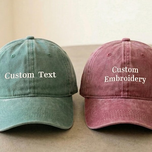 Puede incluir: Dos gorras de béisbol, una verde azulado y otra granate, una al lado de la otra. La gorra verde azulado tiene el texto "Custom Text" bordado en blanco. La gorra granate tiene "Custom Embroidery" bordado en blanco. Ambas gorras tienen visera curva.