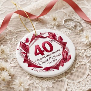 Puede incluir: Un adorno redondo de cerámica blanca con un diseño de cristales geométricos rojos y el texto 40th WEDDING ANNIVERSARY David & Sarah 10.25.1983. El adorno incluye un cordón dorado para colgar y está decorado con detalles de purpurina roja.