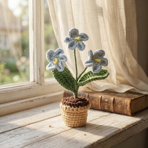 May include: A crocheted flower arrangement featuring three light blue flowers with yellow centers, green leaves, and a brown and tan pot. The handmade flowers sit on a wooden windowsill next to a vintage book.