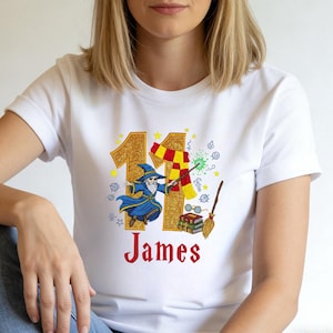 Könnte beinhalten: Weißes T-Shirt mit einem Zauberer-Design, der Zahl 11 in Goldglitter, einem rot-goldenen Schal und dem Namen "James" in Rot. Enthält einen Zauberstab, eine Brille, Bücher und einen Besen.