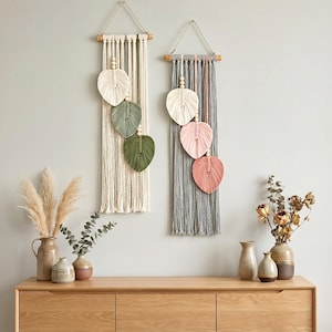 May include: Two macrame wall hangings with leaf designs in cream, gray, pink, and green. Each piece has a wooden dowel and hanging string. Decorative vases with dried plants sit on a wooden cabinet.