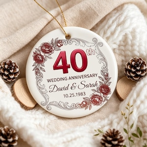 Op de afbeelding: Een rond wit keramieken ornament met een rode 40th tekst, de woorden WEDDING ANNIVERSARY, de namen David & Sarah en de datum 25-10-1983. Het ontwerp bevat een decoratieve grijze krulrand met rode en bruine bloemillustraties als accent.
