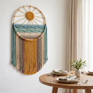 May include: A large, circular macrame wall hanging with a sunburst design. The top section features a woven sun in mustard yellow, with radiating lines. Below, the piece transitions to shades of blue, green, and gold, with long, fringed strands. The piece is mounted on a wooden frame.