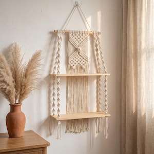 May include: A two-tiered macrame hanging shelf with natural wood shelves and a decorative woven design. The shelf is suspended from a wooden dowel and features intricate knotting details. The neutral color palette complements the bohemian style.