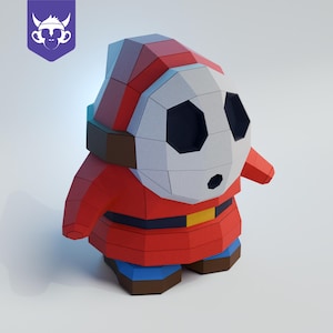 Puede incluir: Una figura de papel de Shy Guy, un personaje de la serie de videojuegos Mario. La figura es principalmente roja con una máscara blanca, agujeros negros para los ojos y un cinturón amarillo. La figura está sobre una superficie blanca.