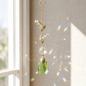 Op de afbeelding: Een hangend zonnevanger-ornament met een goudkleurige metalen steel met witte lelietjes-van-dalen, een heldere gefacetteerde zeshoekige kristallen kraal en een grote, peervormige limoengroene kristallen hanger met een klein gouden bladaccent.