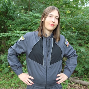 May include: A charcoal grey long-sleeved jacket with a black chest panel and a central zip. The jacket features a rectangular patch with the text 'ATLANTIS' on the right shoulder and a small United States flag patch on the left shoulder.