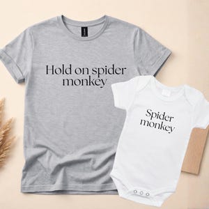 Könnte beinhalten: Ein graues kurzärmeliges T-Shirt mit dem schwarzen Text Hold on spider monkey und ein weißer kurzärmeliger Babybody mit dem schwarzen Text Spider monkey. Beide Kleidungsstücke haben einen einfachen Rundhalsausschnitt.