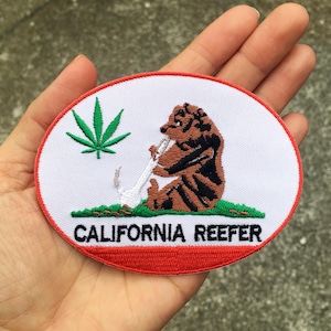 May include: An oval-shaped embroidered patch with a white background and a red border. It features a brown bear sitting on green grass, holding a white smoking device, a green cannabis leaf, and the black text CALIFORNIA REEFER at the bottom.