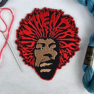 Peut inclure: Écusson brodé noir, rouge et marron représentant un portrait de musicien avec le mot "HEAD" en rouge. L'écusson a une bordure noire. Du fil rouge, une aiguille et du fil à broder bleu sont également visibles.