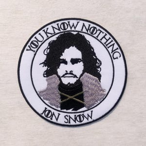 Può includere: Una toppa ricamata circolare con un ritratto in bianco e nero di un uomo con capelli ricci e un cappotto con colletto di pelliccia. Il testo YOU KNOW NOTHING è arcuato sopra e JON SNOW sotto, entrambi in lettere di colore nero.