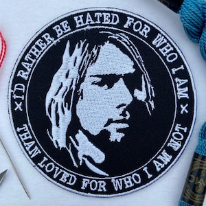 May include: A black and white embroidered patch featuring a portrait of a person with long hair. The patch has the text "I'd rather be hated for who I am than loved for who I am not" in a circular design.