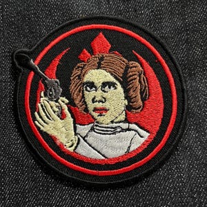 May include: Embroidered patch featuring Princess Leia from Star Wars. The patch has a black background with a red and black circular border. Leia is depicted in a white outfit, holding a blaster. The Star Wars Rebel Alliance symbol is in the background.