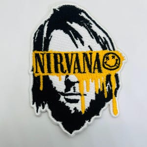 May include: An embroidered patch featuring a black and white stylized portrait of a person with long hair. A horizontal yellow band with a dripping paint effect covers the eyes, displaying the text NIRVANA and a smiley face icon on the right.