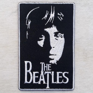 Könnte beinhalten: Schwarz-weißer, gestickter Aufnäher mit einem Porträt eines Mannes und dem Text "THE BEATLES". Der Aufnäher hat eine rechteckige Form mit weißem Rand. Das Design ist eine Hommage an die Kultband.