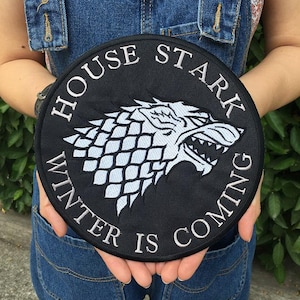 May include: A circular black embroidered patch featuring a white direwolf head emblem in the centre. The text HOUSE STARK is arched across the top, and the text WINTER IS COMING is arched across the bottom in white capital letters.