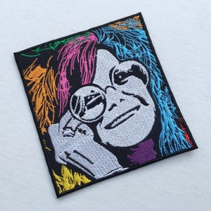 Puede incluir: Parche bordado con un retrato estilizado de una persona con gafas redondas y cabello colorido. El parche tiene un fondo negro con acentos de rosa, azul, amarillo y morado. La cara de la persona es blanca y las gafas son plateadas.