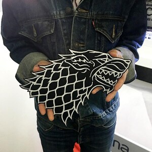 May include: A large, black and white embroidered patch featuring the stylised head of a direwolf in profile. The design showcases intricate white stitching on a black background, depicting the wolf with an open mouth, sharp teeth, and fur.