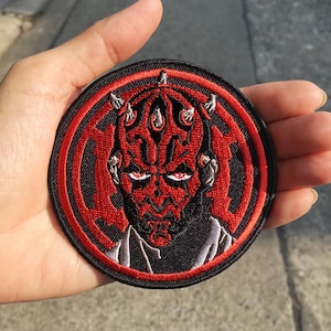 May include: A circular embroidered patch featuring a detailed depiction of a red and black horned character with glowing eyes. The design is set against a red circular background with a black border, showcasing intricate stitching and bold colours.