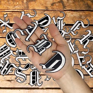 Op de afbeelding: Een verzameling individuele geborduurde patches met letters van het alfabet in een zwart-witte gotische letterstijl. De patches zijn verspreid gerangschikt, waarbij verschillende letters in een handpalm worden gehouden om hun details te tonen.