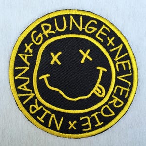 Peut inclure: Un écusson rond noir avec une bordure jaune, représentant un visage souriant. L'écusson porte l'inscription jaune "NIRVANA + GRUNGE + NEVER DIE", entourant le visage souriant avec des yeux barrés et une langue tirée.