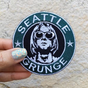 May include: Embroidered patch with a circular design featuring a black and white image of a person wearing sunglasses, surrounded by the words "SEATTLE GRUNGE" in white lettering on a dark green background. Two white stars are on either side of the image.